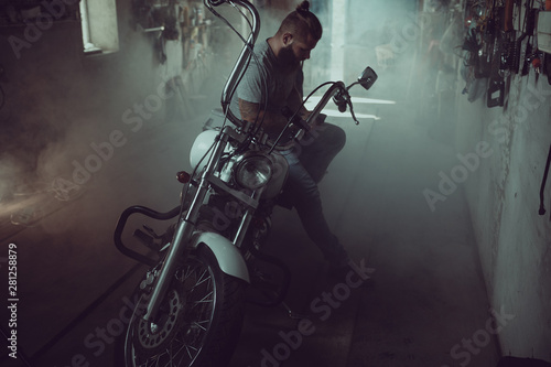 Handsome brutal man with a beard sitting on a motorcycle in his garage, wiping his hands and looking to the side