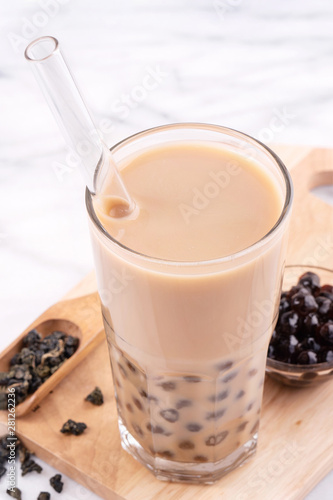 Tapioca pearl ball bubble milk tea, popular Taiwan drink, in drinking glass with straw on marble white table and wooden tray, close up, copy space.