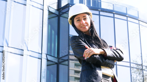 Business woman in suit and helment with crossed arms, looking at camera, smiling. Concept of: Skyscraper background, Communication, Manager, New business.