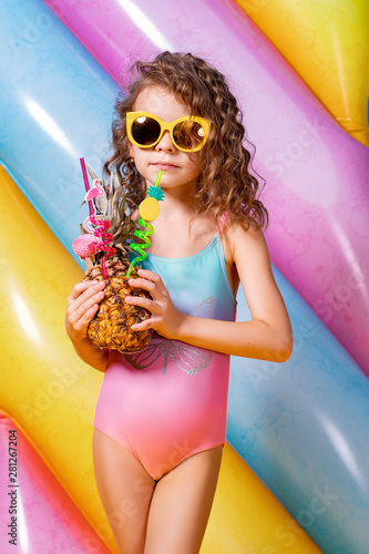 Pretty smiling girl wearing pink and blue swimwear and sunglasses holding pineapple cocktail with colorful straws and showing thumb up on rainbow inflatable mattress background