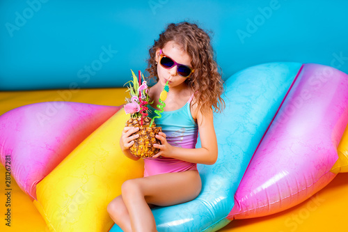 Pretty smiling girl wearing pink and blue swimwear and sunglasses holding pineapple cocktail with colorful straws and showing thumb up on rainbow inflatable mattress background