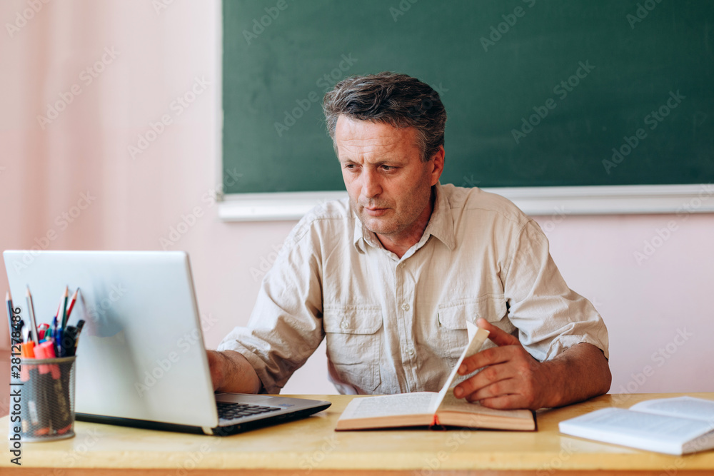 Middle age teacher sitting with open textbook and laptop and working.- Image Middle age teacher sitting with open textbook and laptop and working.- Image