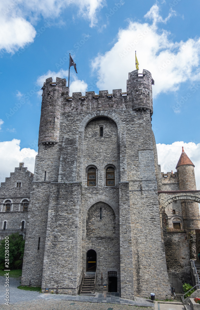 Gent, Flanders, Belgium -  June 21, 2019: Gray stone tower and adjacent buildings of Gravensteen, historic medieval castle of city against blue sky with white clouds. Flags on top, Green foliage.