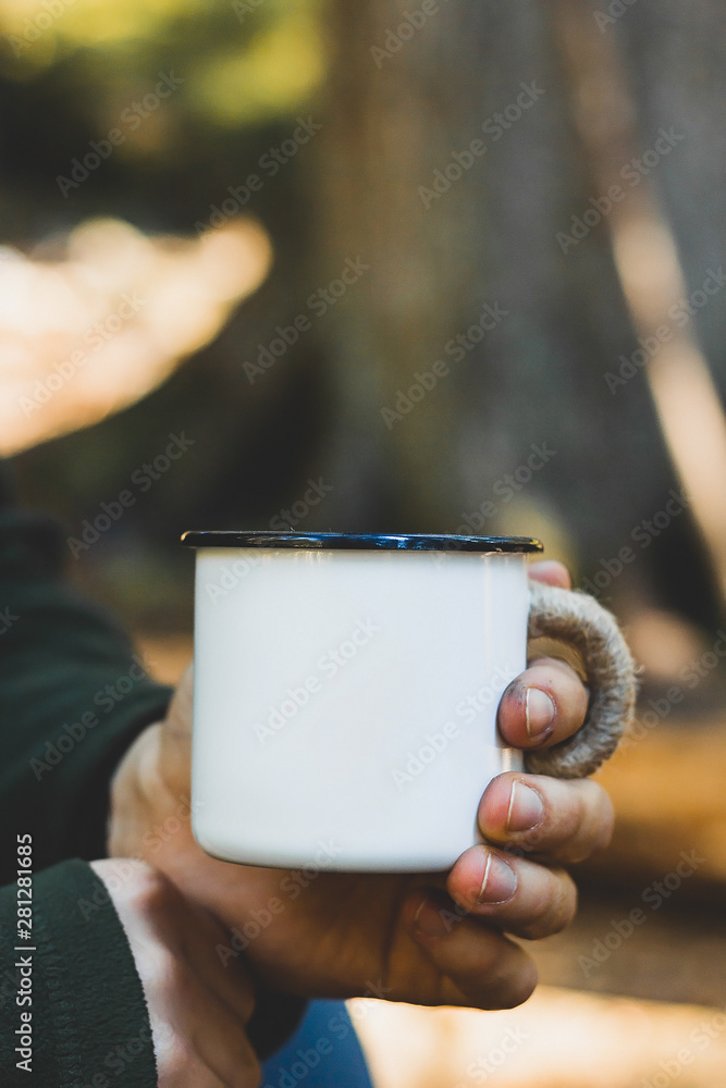 Dirty man's hand holding white enamel cup of hot beverage by an outdoor campfire with a vintage folk edit. Selective focus on mug with blurred background. Close up Dirty man's hand holding white enamel cup of hot beverage by an outdoor campfire with a vintage folk edit. Selective focus on mug with blurred background. Close up