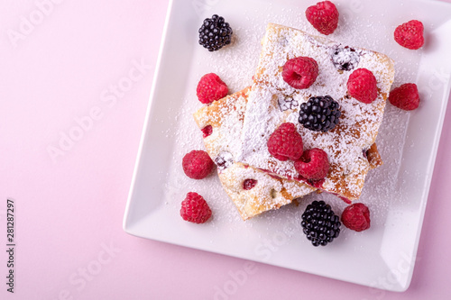 Homemade piece of cake, two pie slices with raspberry and blackberry berries, powdered sugar, in white square plate on pink paper background, top view, flat lay