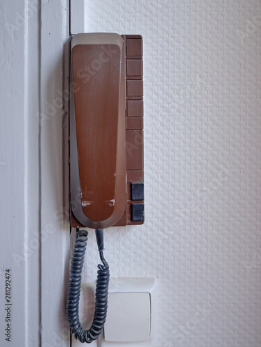 Vintage brown wall phone, hanging off a white textured wall. Phone also has intercom bottoms to let people in front door