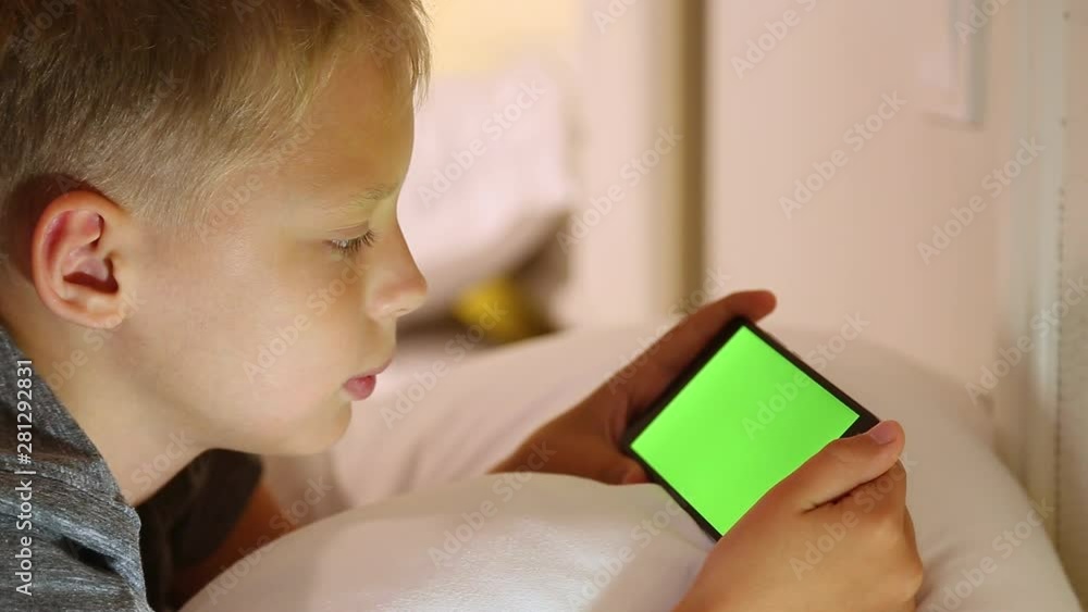 Closeup side view portrait of cute white kid watching video at blank green screen of modern smartphone. Boy laying in bed at home. Real time full hd video footage. Closeup side view portrait of cute white kid watching video at blank green screen of modern smartphone. Boy laying in bed at home. Real time full hd video footage.