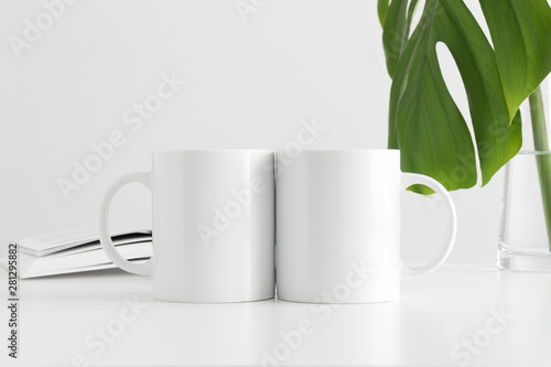 Two white mugs mockup with a monstera leaf in a vase and a book on a white table.