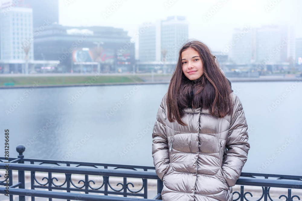 young hipster girl is standing in the park in a warm coat leaning on the railing of the bridge, hands in jacket pockets, looking at the camera, river background