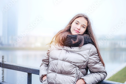 modern young girl teenager stands on the bridge leaning on the railing hiding her hands in the pockets of a warm jacket