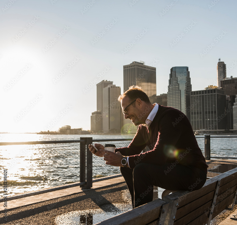 USA, Brooklyn, smiling man with coffee to go sitting on bench looking at smartphone USA, Brooklyn, smiling man with coffee to go sitting on bench looking at smartphone