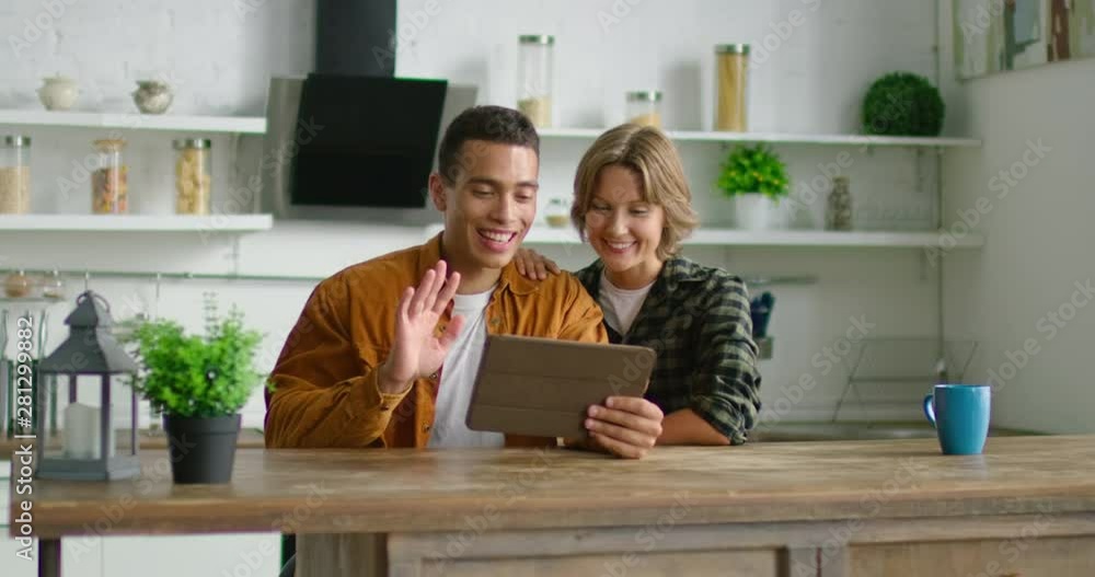 Latin man and white woman talking with freinds, family via video application on tablet computer, waving hello, smiling, sitting at table in kitchen, having joyful time at home. 4K, shot on RED camera. Latin man and white woman talking with freinds, family via video application on tablet computer, waving hello, smiling, sitting at table in kitchen, having joyful time at home. 4K, shot on RED camera.