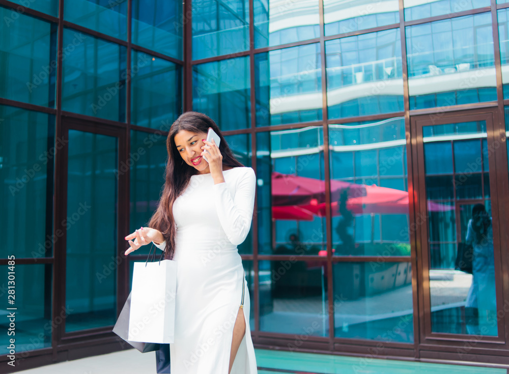 Young afro woman in white dress with paper shopping bags talking on the phone outdoors against the background of a business building