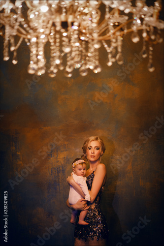 Tender photo of beautiful mother in fashion dress with luxurious blond hair posing with her cute little baby girl in studio. Mother and little daughter like a queen and princess