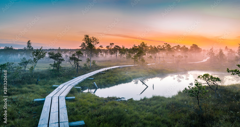 Warmly colored sunrise over a foggy swamp. Aerial view of stunning landscape at peat bog at Kemeri National park in Latvia. Wooden trail leading along the lake surrounded by pounds and forest.