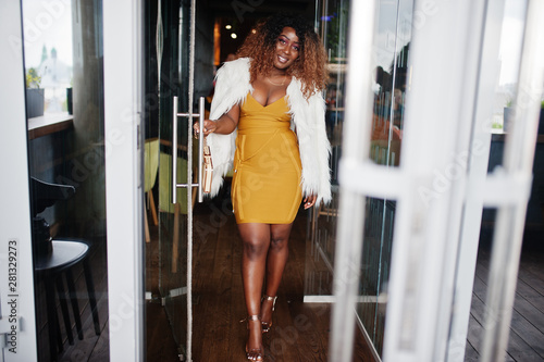 Glamour african american woman in yellow dress and white woolen cape with handbag posed at glass door entrance of restaurant.