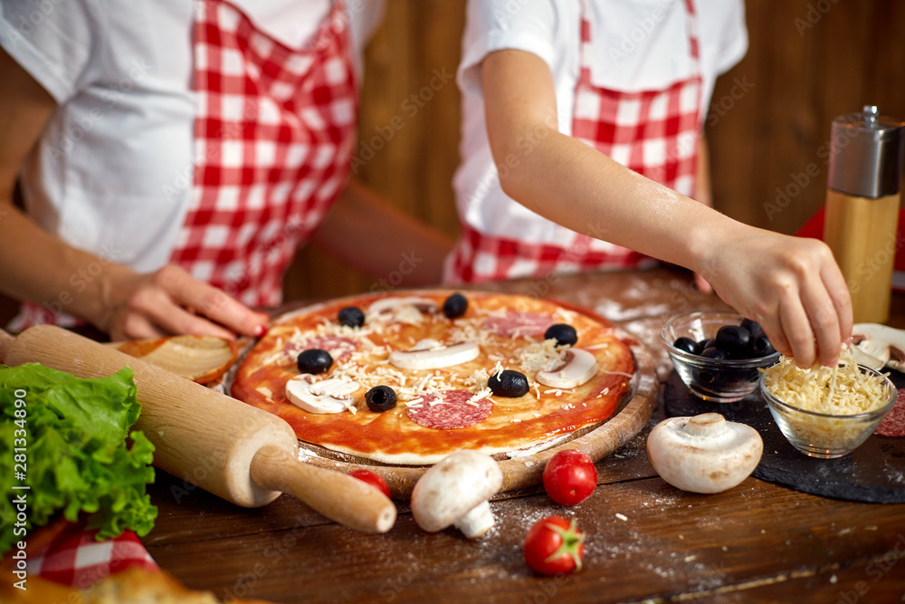 mother and daughter wearing white T-shirts and checkered aprons cooking pizza together and decorating with mushrooms on table filled with ingredients in stylish wooden kitchen