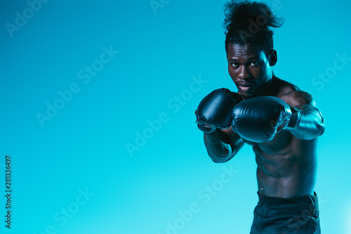 shirtless african american sportsman in boxing gloves looking at camera on blue background