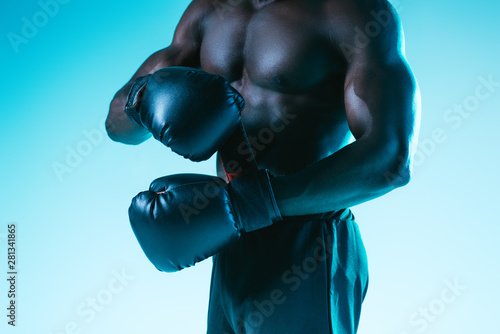 cropped view of shirtless, muscular african american sportsman in boxing gloves on blue background