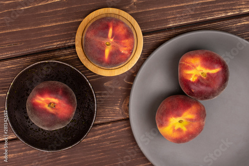 Group of four whole fresh red peach on a round bamboo coaster in a dark ceramic bowl on a gray ceramic plate flatlay on brown wood
