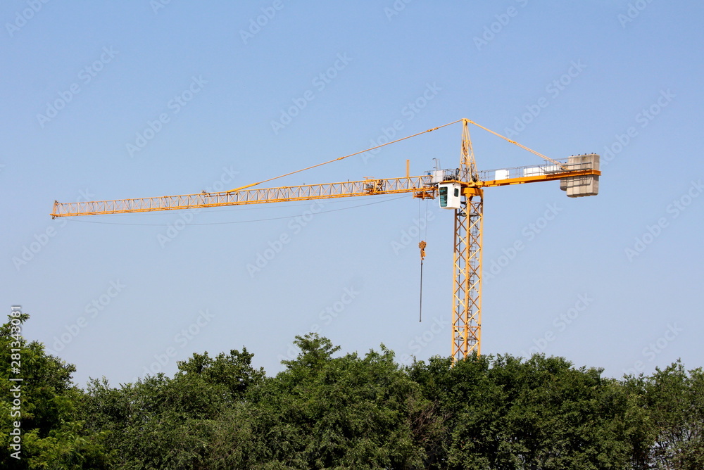 Construction industry tower crane or TC rising high over construction site surrounded with dense forest with clear blue sky in background Construction industry tower crane or TC rising high over construction site surrounded with dense forest with clear blue sky in background