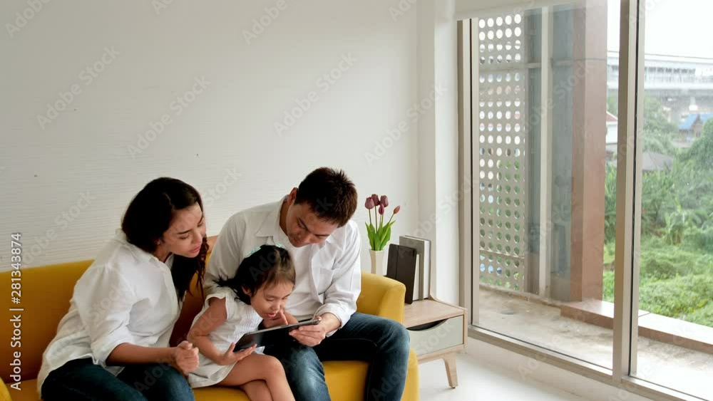 Asian family happy on sofa in the house with sunlight from the window. Father, mother and daughter, aged three, watching online media with a digital tablet. Family leisure activities, education Asian family happy on sofa in the house with sunlight from the window. Father, mother and daughter, aged three, watching online media with a digital tablet. Family leisure activities, education
