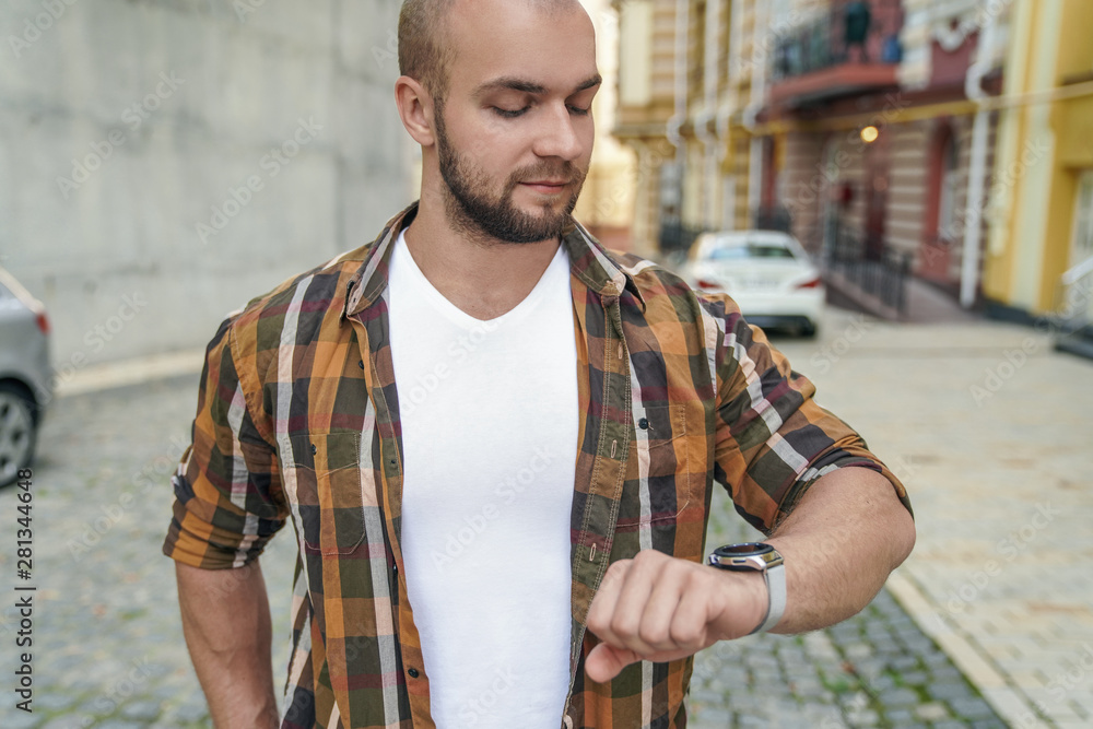 Obraz premium young good looking bold bearded guy standing outdoors near his house checking time making sure he is not late. smiling. wearing yellow shirt and jeans.