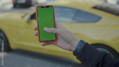 Close-up young rich businessman using mobile phone tapping on screen standing near yellow expensiv sports car in the street. Mockup green screen blank smartphone. Luxurious life. photo