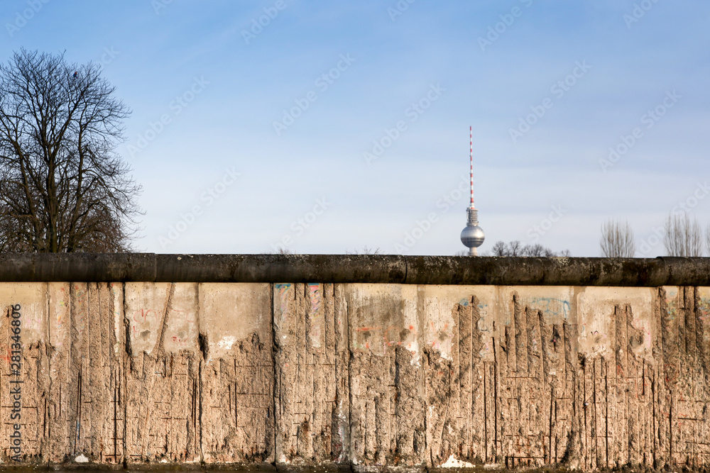 Berlin Wall original weathered section damaged with exposed iron bars partly covering the TV tower (Berliner Fernsehturm) far in the horizon.