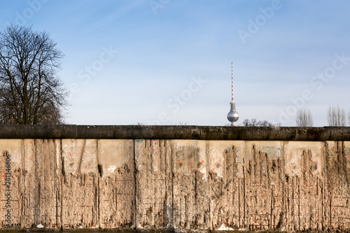 Photography Berlin Wall original weathered section damaged with exposed iron bars partly covering the TV tower (Berliner Fernsehturm) far in the horizon
