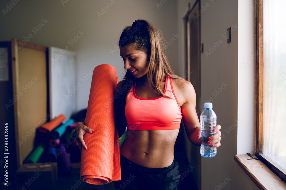 Muscular sporty fit sportswoman holding bottle of water and yoga mat by the window at the gym studio going to the training workout