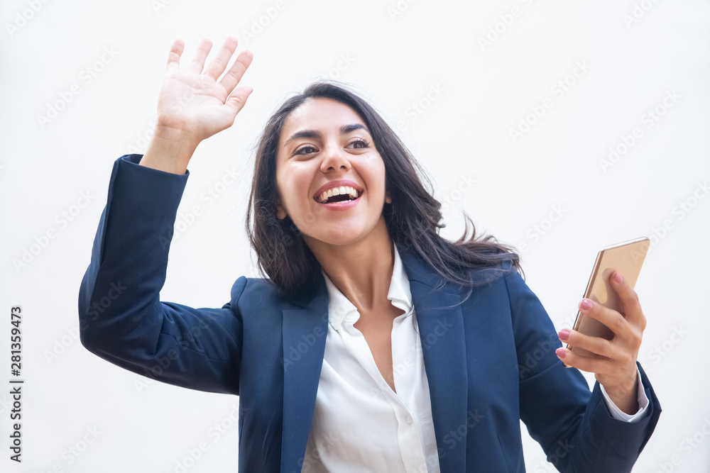 Happy businesswoman with smartphone greeting someone. Beautiful young woman in formal wear holding mobile phone and waving hand isolated on grey background. Technology concept Happy businesswoman with smartphone greeting someone. Beautiful young woman in formal wear holding mobile phone and waving hand isolated on grey background. Technology concept