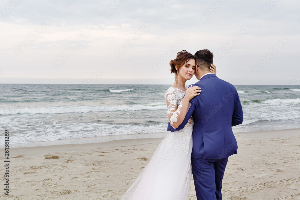 newlyweds walking along the sea beach at the wedding. The groom hugs the bride newlyweds walking along the sea beach at the wedding. The groom hugs the bride