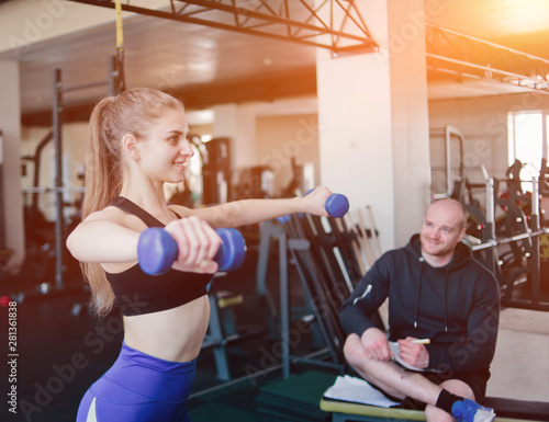 Fitness instructor supervises and notes in notebook results of training young athletic blonde performing exercises with dumbbells in her hands at gym