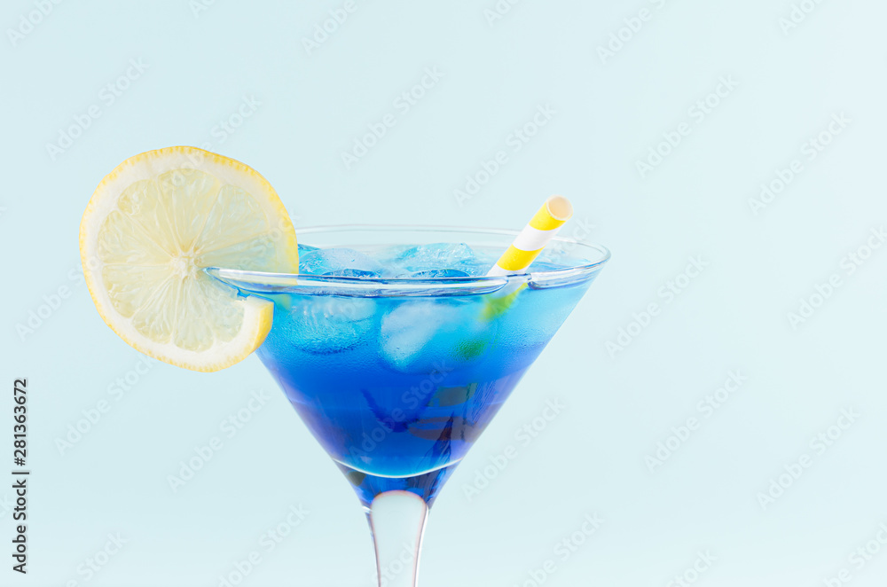 Cool summer blue beverage margarita for party with ice cubes, lemon slice, yellow straw on white wood board and mint color wall, top part, details. Cool summer blue beverage margarita for party with ice cubes, lemon slice, yellow straw on white wood board and mint color wall, top part, details.