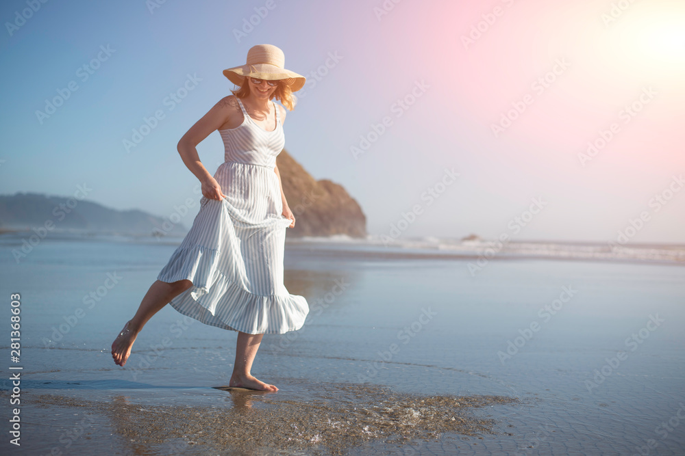 A girl in a hat at sunset near the ocean splashes water with splashes. A woman's style and fashion, relaxing on the beach, walking, with space