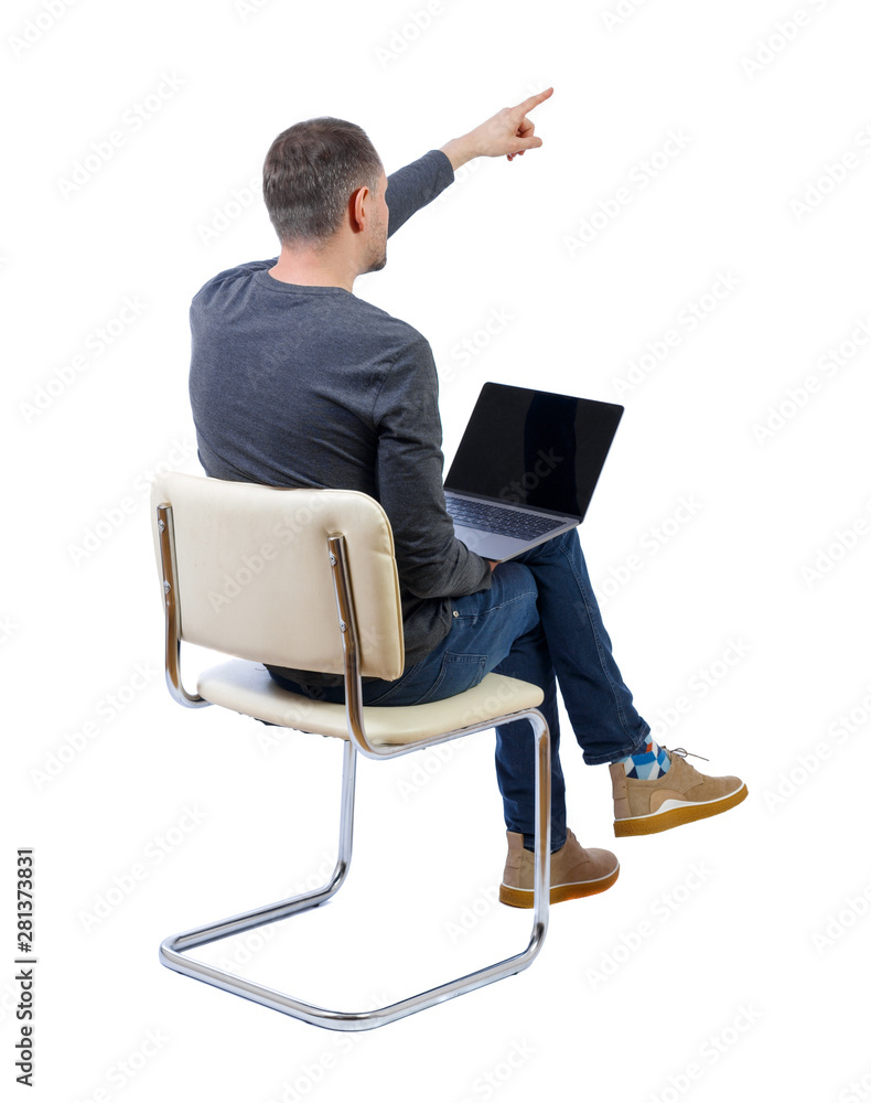 Back view of a man who sits on a chair with a laptop and points with his hand forward.