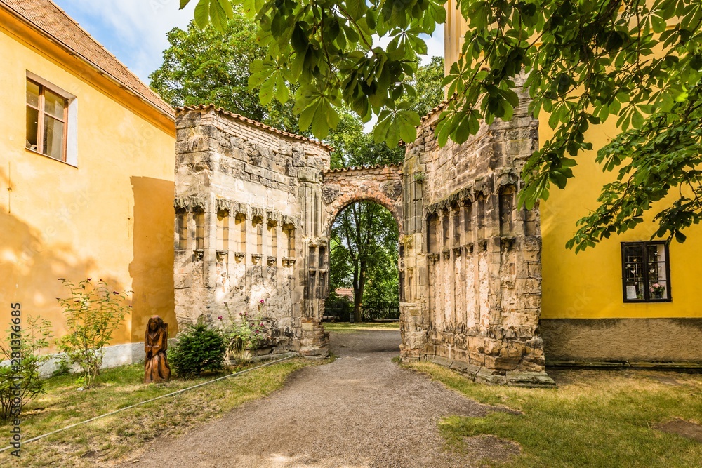 Panensky Tynec, Czech Republic - July 15 2019: Stone gateway of the unfinished Gothic church of the Virgin Mary from 14th century. Yellow buildings. Sunny summer day. Green chestnut tree in foreground