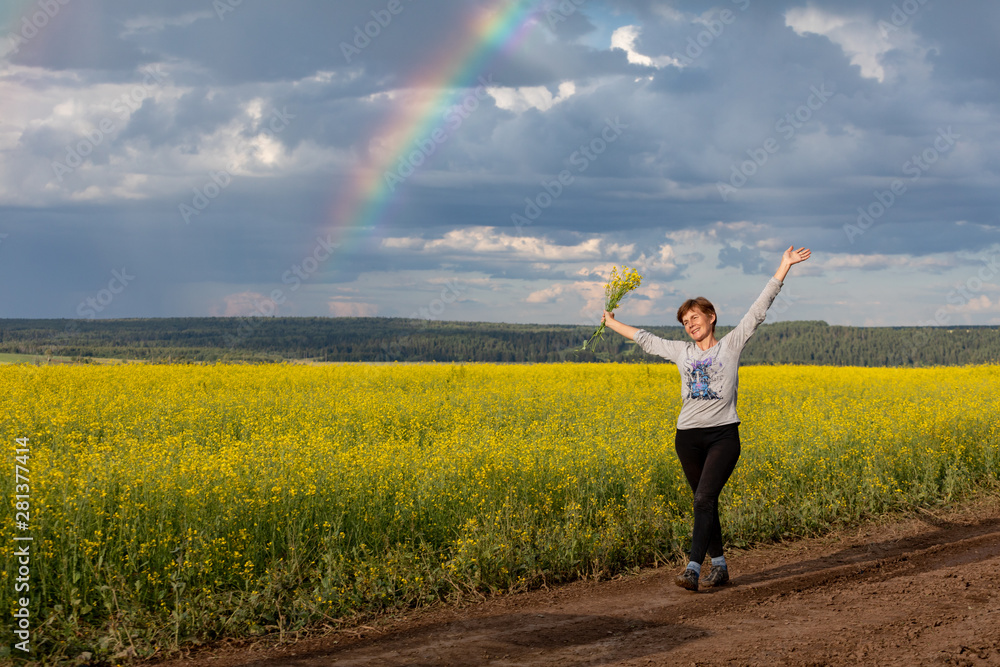 Fototapeta premium A girl is walking along a rural road through a blooming yellow field. Sunny summer day