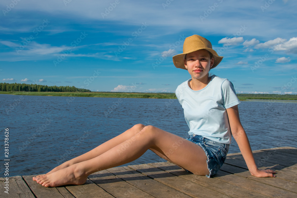 the girl in the hat sits on the pier, against the blue sky and the lake, gets a sun bath