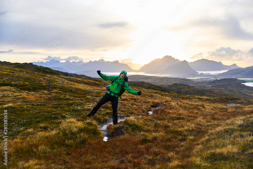 Young man hiking and cheering on Lofoten Islands in Norway with a sunset behind the mountains.