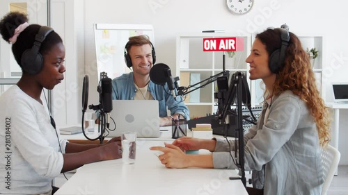 Medium shot of three diverse radio hosts sitting at microphones at table in studio and having pleasant talk on air photo