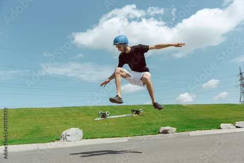 Skateboarder doing a trick at the city's street in sunny day. Young man in equipment riding and longboarding near by meadow in action. Concept of leisure activity, sport, extreme, hobby and motion.