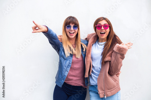 Two young pretty women outdoor and  one of them points at something spring, fashion, accessories and people concept - happy women in sunglasses pointing finger at white wall background 