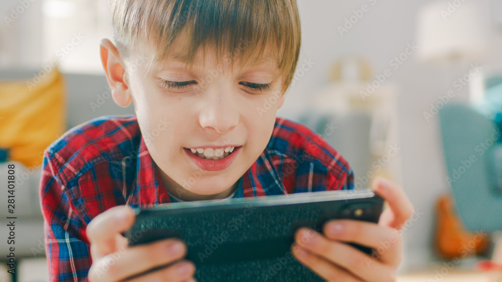 Close-up Portrait of a Smart Little Boy Laying on a Carpet Plays in Video Game on His Smartphone, Holds Mobile Phone in Horizontal Landscape Mode. Child Has Fun Playing Videogame in Sunny Living Room. Close-up Portrait of a Smart Little Boy Laying on a Carpet Plays in Video Game on His Smartphone, Holds Mobile Phone in Horizontal Landscape Mode. Child Has Fun Playing Videogame in Sunny Living Room.