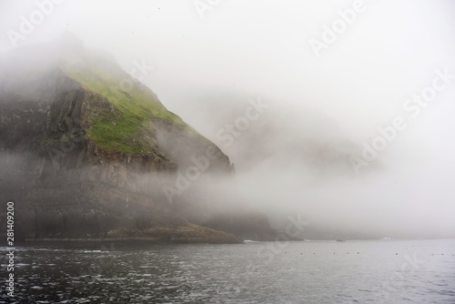 Fantastic morning scene of Mykines island. Green cliffs above the ocean and thick morning fog over the ocean.