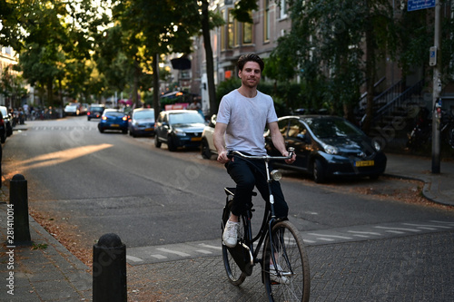 Spontaneous young Dutch man smiling and riding his bicycle through an old and cosy street in Amsterdam, the Netherlands