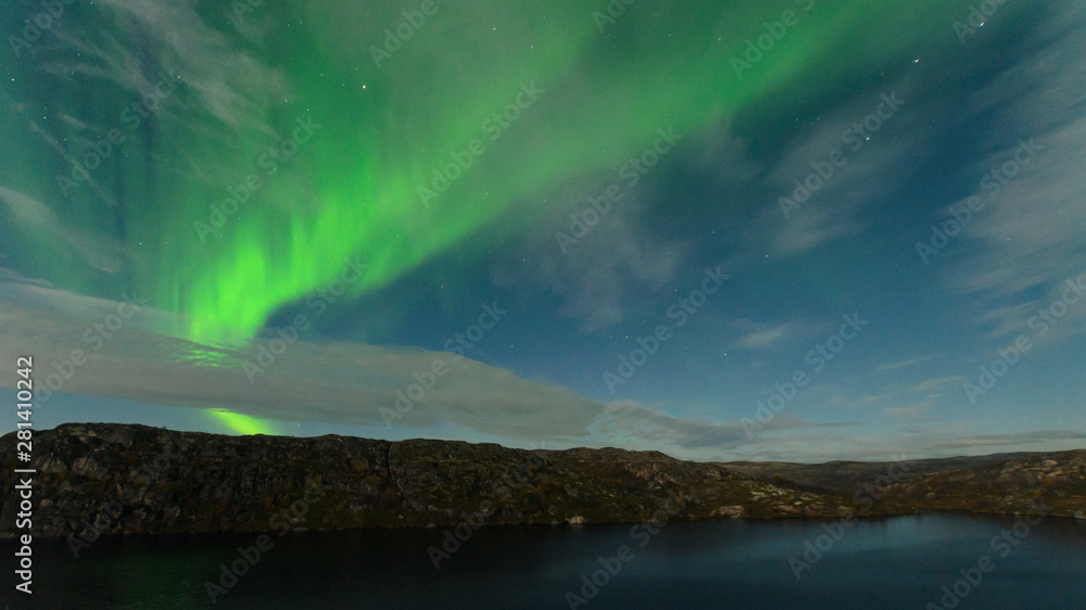 Northern lights, aurora in the sky above the hills and cliffs and reflected in the lake.