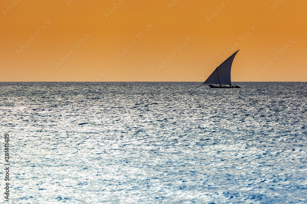 Traditional dhow sailing vessel used to transport people goods and commodities silhouetted against a warm sky and blue horizon