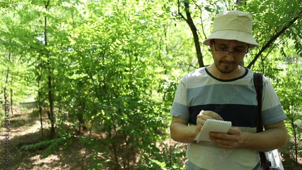 Cute caucasian biologist with glasses, Panama and stubble, stands in the green forest, and takes notes in his notebook, close-up Cute caucasian biologist with glasses, Panama and stubble, stands in the green forest, and takes notes in his notebook, close-up
